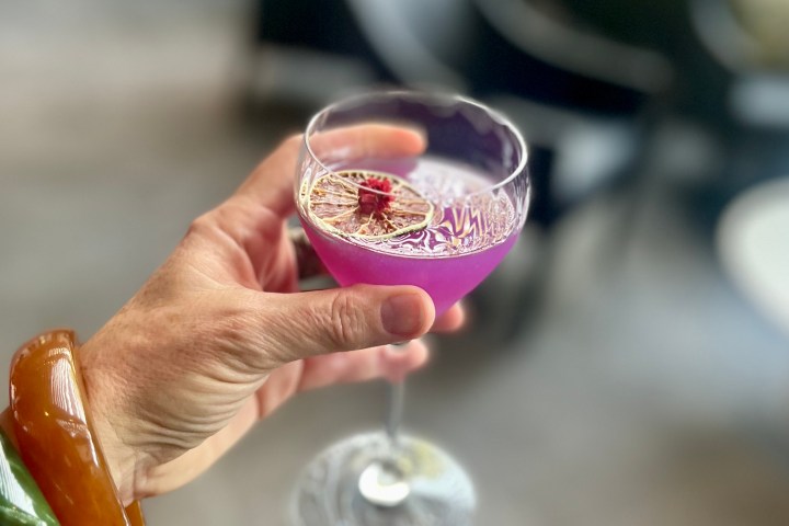 Hand holding a purple cocktail with a flower garnish in a glass.
